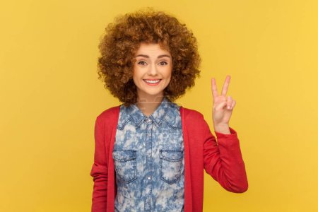 Photo for Portrait of happy optimistic woman with Afro hairstyle showing two fingers V sign, looking at camera with toothy smile, victory gesture. Indoor studio shot isolated on yellow background. - Royalty Free Image
