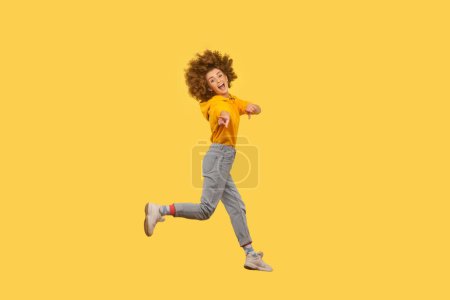 Retrato de mujer emocionada satisfecha con peinado afro saltando alto, apuntándote, eligiéndote, usando sudadera con capucha estilo casual. Estudio interior plano aislado sobre fondo amarillo.