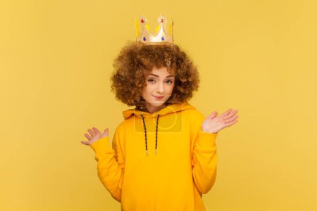 Portrait of self- confident egoistic beautiful woman with Afro hairstyle in golden crown, pretended to be queen, wearing casual style hoodie. Indoor studio shot isolated on yellow background.