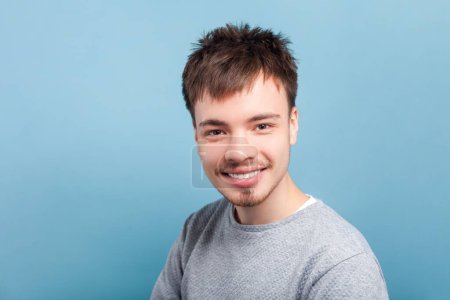 Portrait of attractive smiling happy positive man wearing gray jumper looking at camera being in good mood optimistic emotions. Indoor studio shot isolated on blue background.