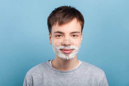 Photo for Portrait of smiling attractive man wearing gray jumper standing with face shaving foam doing morning skin care procedures. Indoor studio shot isolated on blue background. - Royalty Free Image