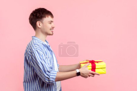 Photo for Side view portrait of smiling happy young man wearing shirt standing holding yellow present box congratulating with birthday. Indoor studio shot isolated on pink background. - Royalty Free Image