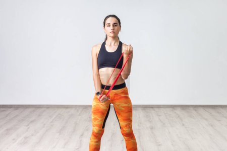 Portrait of powerful sporty woman wearing black tank top and orange leggings, doing exercise for her arms with elastic band. Full length indoor shot against white wall.