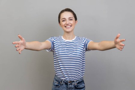 Komm in meine Arme. Porträt einer glücklichen positiven Frau in gestreiftem T-Shirt, die in die Kamera streckt und die Arme ausstreckt, um dich zu umarmen. Indoor Studio isoliert auf grauem Hintergrund gedreht.