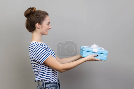 Photo for Side view of woman wearing striped T-shirt holding out present box, giving gift, looking ahead with optimistic expression, greeting. Indoor studio shot isolated on gray background. - Royalty Free Image