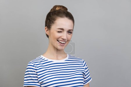 Photo for Portrait of cheerful woman wearing striped T-shirt standing and winking playfully, having positive expression, looking at camera. Indoor studio shot isolated on gray background. - Royalty Free Image