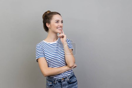 Photo for Portrait of smiling woman wearing striped T-shirt standing looking away, keeping finger on cheek, dreaming, making wish, love. Indoor studio shot isolated on gray background. - Royalty Free Image