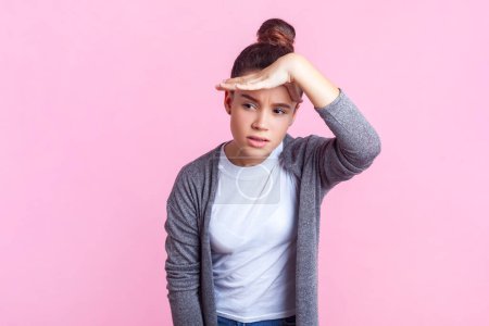 Portrait d'adolescente concentrée sérieuse avec une coiffure de chignon dans des vêtements décontractés debout regardant loin avec une expression concentrée. Studio intérieur tourné isolé sur fond rose.