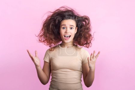 Portrait of amazed surprised angry teenage girl with wavy tousled hair in beige T- shirt standing asking why with raised arms. Indoor studio shot isolated on pink background.