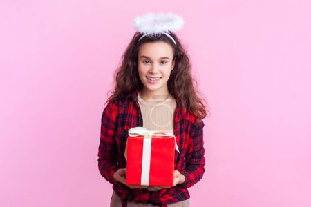 Portrait of pretty pleased teenage girl with wavy hair in red checkered shirt and nimb over head standing giving present box charity. Indoor studio shot isolated on pink background.