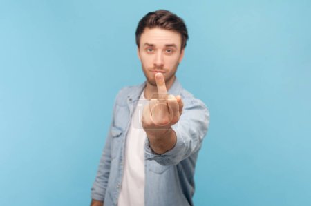 Portrait of unshaven man rejecting communication, showing middle finger to express disrespect and hate, vulgar gesture, wearing denim shirt. Indoor shot isolated on blue background.
