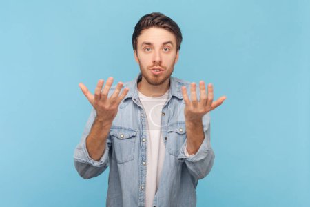 Portrait of confused or shocked handsome unshaven man standing with raised arms and looking at camera asking expression, wearing denim shirt. Indoor shot isolated on blue background.