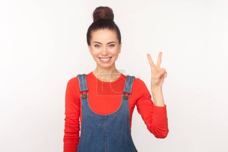 Photo for Satisfied smiling attractive woman with hair bun looking at camera with happy expression, showing v sign, victory gesture, wearing denim overalls. Indoor studio shot isolated on white background - Royalty Free Image
