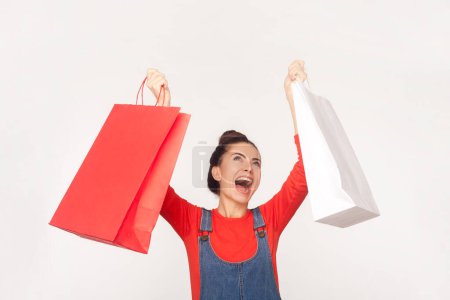 Portrait of emotional happy satisfied woman with hair bun holding shopping bags end rejoicing her purchases, dancing, wearing denim overalls. Indoor studio shot isolated on white background