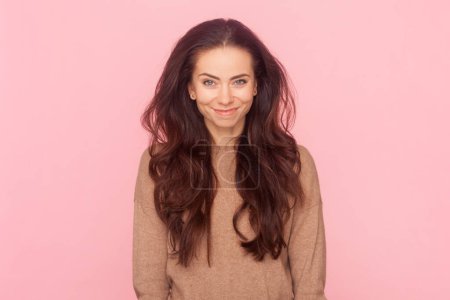 Téléchargez les photos : Portrait de femme attrayante positive souriante aux cheveux ondulés, regardant la caméra avec un visage rusé, étant de bonne humeur, portant un pull marron. Studio intérieur tourné isolé sur fond rose - en image libre de droit
