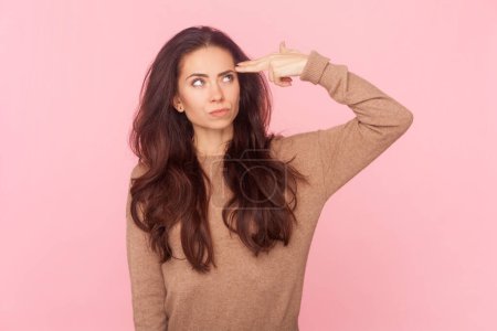 Portrait of sad despair stressed woman with wavy hair, making gun suicide gesture, having lots problems, being tired, wearing wearing brown pullover. Indoor studio shot isolated on pink background