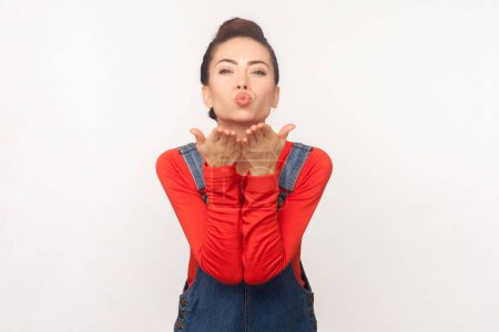 Photo for Woman with hair bun blows air kiss keeps lips folded palm towards mouth sends mwah coquettish wants to cheer up her partner, wearing denim overalls. Indoor studio shot isolated on white background - Royalty Free Image