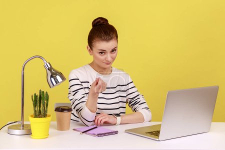 Une jeune femme gestionnaire positive assise sur le lieu de travail avec un ordinateur portable et montrant un geste d'argent, demandant un paiement, planifiant le revenu de l'entreprise. Studio intérieur tourné isolé sur fond jaune.