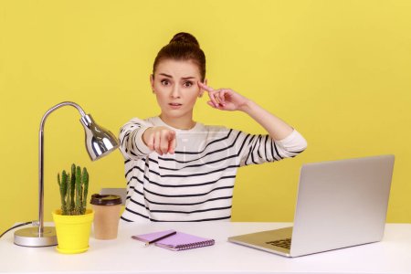 Foto de Woman with sarcastic look sitting at workplace and making stupid gesture, pointing to camera, teasing and accusing senseless dumb mind. Indoor studio studio shot isolated on yellow background. - Imagen libre de derechos