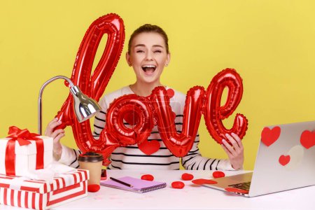 Photo for Satisfied excited woman holding love word of foil balloons, expressing romantic emotions and feeling, sitting on workplace with laptop. Indoor studio studio shot isolated on yellow background. - Royalty Free Image