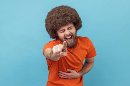 Porträt eines glücklichen Mannes mit Afro-Frisur, der ein orangefarbenes T-Shirt trägt, mit dem Finger auf dich zeigt und aufrichtig lachend die Hand auf dem Bauch hält. Spaß haben. Indoor Studio aufgenommen isoliert auf blauem Hintergrund.