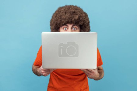 Photo for Portrait of surprised amazed man with Afro hairstyle wearing orange T-shirt covering half of his face with laptop, looking at camera with big eyes. Indoor studio shot isolated on blue background. - Royalty Free Image