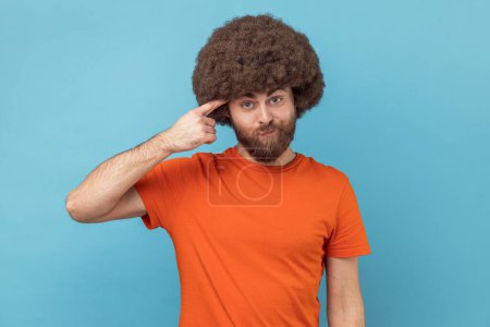 Photo for Portrait of man with Afro hairstyle in orange T-shirt holding finger against his temple, making crazy stupid gesture, blaming in senseless dumb talk. Indoor studio shot isolated on blue background. - Royalty Free Image