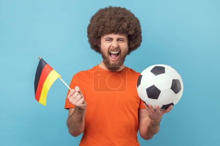Portrait of crazy man with Afro hairstyle holding flag of germany and soccer black and white classic ball and watching match, cheering. Indoor studio shot isolated on blue background.