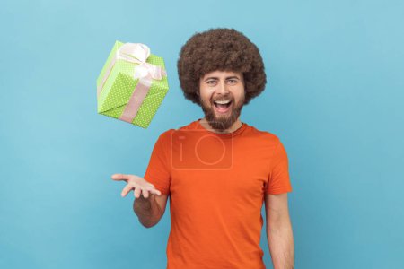 Téléchargez les photos : Portrait d'homme joyeux et optimiste avec une coiffure afro portant un T-shirt orange jetant une boîte cadeau, s'amusant à la fête d'anniversaire. Studio intérieur tourné isolé sur fond bleu. - en image libre de droit