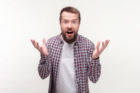 Portrait of angry annoyed bearded man standing with raised arms arguing asking what why, wearing casual checkered shirt. Indoor studio shot isolated on gray background.