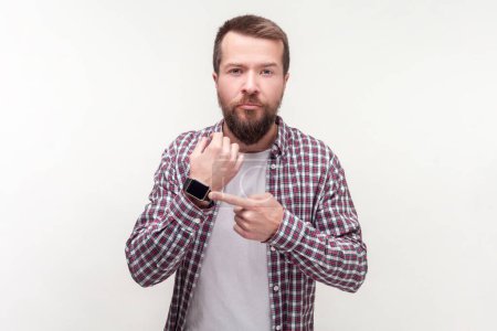 Téléchargez les photos : Portrait d'un homme barbu sérieux et confiant pointant sa montre-bracelet en regardant la date limite de synchronisation de la caméra, portant une chemise à carreaux décontractée. Studio intérieur tourné isolé sur fond gris. - en image libre de droit
