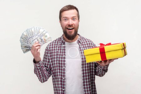 Photo for Portrait of amazed excited handsome bearded man holding dollar banknotes and yellow present box, wearing casual checkered shirt. Indoor studio shot isolated on gray background. - Royalty Free Image