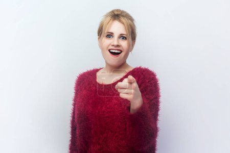 Photo for Portrait of laughing young blonde woman standing pointing at camera being amazed and excited, wearing red fluffy jumper. Indoor studio shot isolated on light gray background. - Royalty Free Image