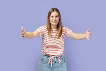 Come into my arms, free hugs. Portrait of adorable hospitable blond woman wearing striped T-shirt smiling and reaching out hands, going to embrace. Indoor studio shot isolated on purple background.