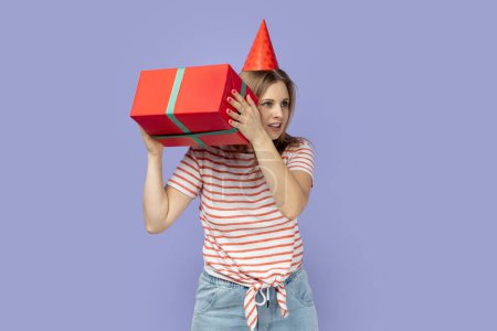 Photo for Portrait of curious blond woman wearing striped T-shirt and party cone shaking present box, thinking what inside, looking away with smile. Indoor studio shot isolated on purple background. - Royalty Free Image