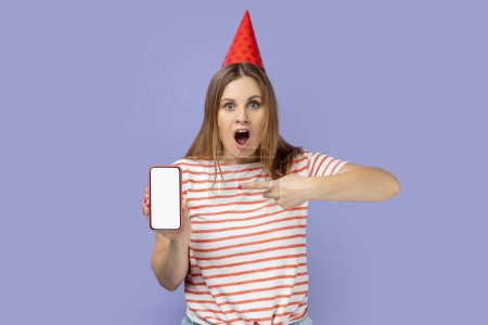Téléchargez les photos : Choquée femme blonde étonnée portant un cône de fête célébrant son anniversaire, montrant téléphone portable avec écran blanc pour la publicité. Studio intérieur tourné isolé sur fond violet. - en image libre de droit