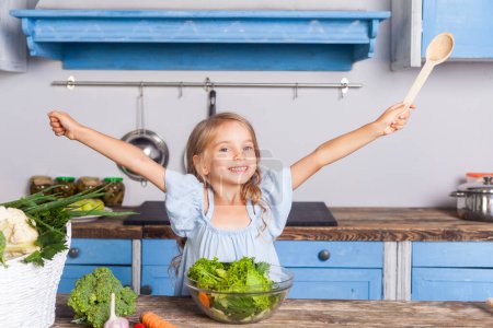 Téléchargez les photos : Aliments végétariens, vitamines et régime alimentaire. Portrait de petite fille tenant une cuillère en bois et souriant regardant la caméra, debout dans la cuisine moderne préparant une salade de légumes, cuisinant un petit déjeuner sain. - en image libre de droit