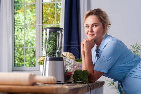 Photo for Attractive blonde woman posing near blender to make healthy smoothie for breakfast, cooking vegetables salad and organic detox meal, posing in modern kitchen near window. - Royalty Free Image