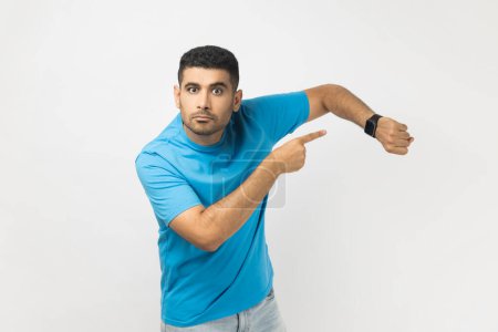 Photo for Time to go, hurry up. Portrait of unshaven man in blue T- shirt standing pointing at wrist, makes time gesture, shows we should do everything quickly. Indoor studio shot isolated on gray background. - Royalty Free Image