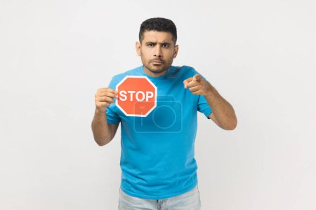 Photo for Portrait of serious angry unshaven man wearing blue T- shirt standing holding red ban symbol and pointing at camera, trying to stop you. Indoor studio shot isolated on gray background. - Royalty Free Image