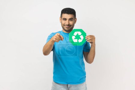 Photo for Portrait of unshaven man wearing blue T- shirt standing holding green recycling sign, pointing to camera, calls on to save our planet from pollution. Indoor studio shot isolated on gray background. - Royalty Free Image