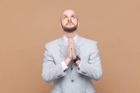 Portrait of attractive bald bearded man raising hands in prayer, looking up with hopeful pleading eyes, praying, wearing gray jacket. Indoor studio shot isolated on brown background.