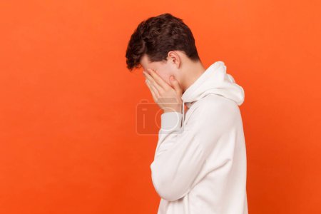 Photo for Portrait of depressed brunette young man wearing white hoodie hiding face in hand, looking desperate, is about to cry, empty copy space for text. Indoor studio shot isolated on orange background. - Royalty Free Image
