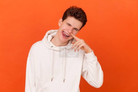 Portrait of uncultured young man wearing white hoodie putting finger inside nose and drilling, fooling around, showing his bad manners. Indoor studio shot isolated on orange background.