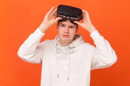 Photo for Portrait of young man wearing white hoodie standing in virtual reality goggles, raising VR headset and looking at camera with serious expression. Indoor studio shot isolated on orange background. - Royalty Free Image