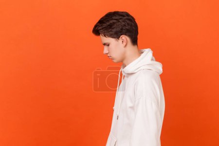 Photo for Portrait of depressed brunette upset young man wearing white hoodie standing with head down, expressing sorrow and sadness. Indoor studio shot isolated on orange background. - Royalty Free Image