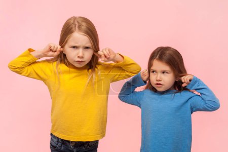 Photo for Portrait of two serious irritated frustrated little girls covering ears with fingers, trying to avoid loud sounds, being in bad mood. Indoor studio shot isolated on pink background. - Royalty Free Image