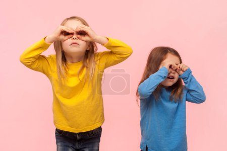 Retrato de dos graciosas niñas curiosas de pie haciendo gestos binoculares, mirando hacia otro lado, esperando algo, jugando, divirtiéndose. Estudio interior plano aislado sobre fondo rosa.