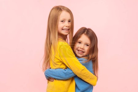 Téléchargez les photos : Portrait de mignon extrêmement heureux souriant deux petites filles debout se serrant dans leurs bras, exprimant l'amour, les soins et le soutien, étant satisfait. Studio intérieur tourné isolé sur fond rose. - en image libre de droit