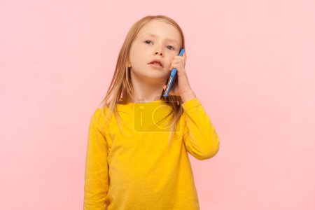 Téléchargez les photos : Portrait d'une petite fille blonde sérieuse cool debout avec un téléphone intelligent, parlant sur un téléphone portable avec un regard confiant, portant un pull jaune. Studio intérieur tourné isolé sur fond rose. - en image libre de droit